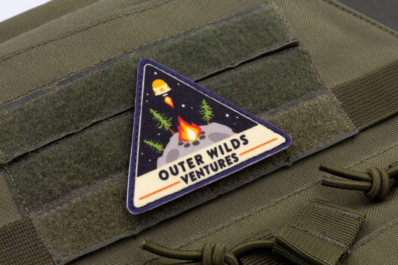 Może przedstawiać: Tr&oacute;jkątna naszywka z napisem "OUTER WILDS VENTURES" przedstawia rakietę, ognisko i drzewa na tle gwiaździstego nieba. Naszywka jest obramowana w kolorze niebieskim i pomarańczowym, na zielonym tle materiału.