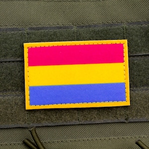 Op de afbeelding: Een rechthoekige patch met een ontwerp van roze, gele en blauwe horizontale strepen. De patch is omrand met gele stiksels en is bevestigd op een groene stoffen achtergrond. Het is waarschijnlijk een embleem of vlag.