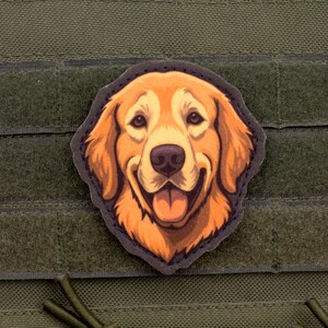 Puede incluir: Un parche con la cara de un perro Golden Retriever sonriente con pelaje naranja y marrón, una nariz negra y una lengua rosa. El parche está sobre un fondo verde con un cierre de velcro.