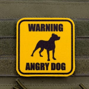 May include: A yellow patch with a black border and the words "WARNING ANGRY DOG" in black. A black silhouette of a dog is in the center. The patch is attached to a green fabric.