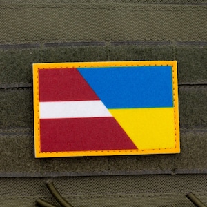 May include: A rectangular patch featuring the flags of Latvia and Ukraine. The Latvian flag is red with a white stripe, and the Ukrainian flag is blue and yellow. The patch has a yellow border and is attached to a green fabric.