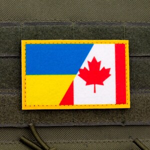 Puede incluir: Un parche rectangular con las banderas de Ucrania y Canadá. La bandera ucraniana es azul y amarilla, mientras que la bandera canadiense presenta una hoja de arce roja sobre fondo blanco. El parche tiene un borde amarillo y está sobre un fondo verde.