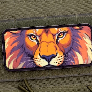 Puede incluir: Un parche rectangular con la cara estilizada de un león. El león tiene pelaje naranja, melena morada y ojos amarillos. El parche está sobre un fondo verde, con un cierre de velcro.
