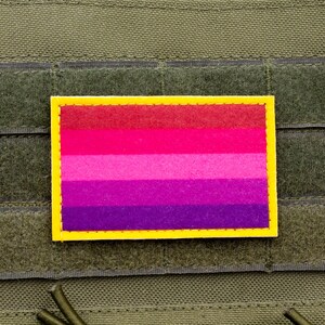 May include: A rectangular patch with a yellow border and a horizontal striped design in shades of red, pink, and purple. The patch is affixed to a green fabric surface with a hook and loop fastener.