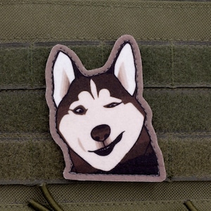 Peut inclure: Écusson de chien husky souriant, avec un design blanc, marron et noir. Le patch rectangulaire a une bordure grise et est fixé sur un fond en tissu vert. Le visage du chien est au centre.