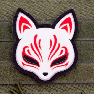 May include: A white and red fox mask patch with black accents, set against a green background. The patch features a stylized fox face design with red markings and black eye shapes. The patch is attached to a green fabric.