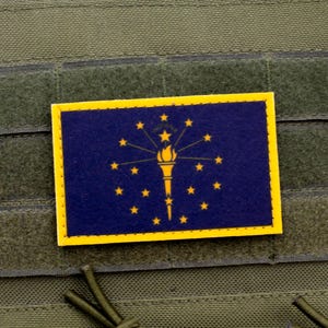 May include: A yellow and blue patch with the state flag of Indiana. The flag features a torch and 19 stars on a blue background.