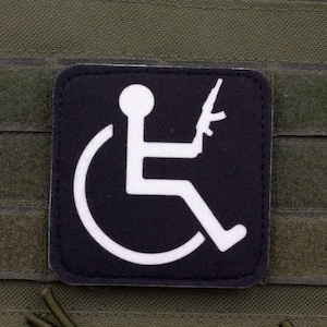 Puede incluir: Un parche cuadrado negro con un gráfico blanco de una persona en silla de ruedas sosteniendo un rifle. El parche tiene esquinas redondeadas y está sujeto a una superficie de tela verde oliva con correas horizontales.