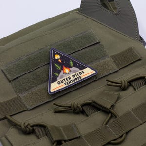 Może przedstawiać: Zielona kamizelka taktyczna z wieloma paskami i tr&oacute;jkątną naszywką. Naszywka przedstawia rakietę, ognisko i tekst "OUTER WILDS VENTURES". Kamizelka posiada zapięcia na rzepy.