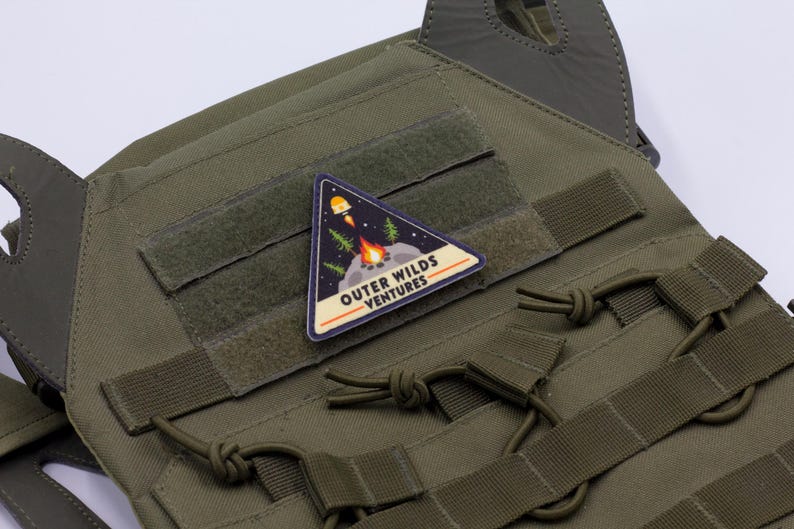 Może przedstawiać: Zielona kamizelka taktyczna z wieloma paskami i tr&oacute;jkątną naszywką. Naszywka przedstawia rakietę, ognisko i tekst "OUTER WILDS VENTURES". Kamizelka posiada zapięcia na rzepy.