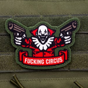 May include: A green patch featuring a clown with two guns, red and white details, and the text "FUCKING CIRCUS" on a red banner. The patch is set against a green background.