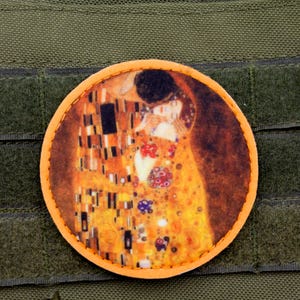 Op de afbeelding: Ronde patch met Gustav Klimt's "De Kus" in goud, oranje en zwart. De patch is geplaatst op een groene achtergrond met klittenbandsluitingen, geschikt voor tactische uitrusting of kleding.