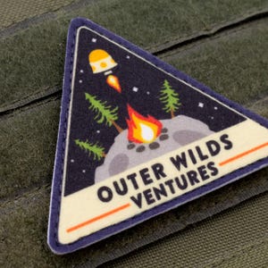 Może przedstawiać: Tr&oacute;jkątna naszywka z granatowym tłem przedstawiająca rakietę, ognisko i drzewa. Na naszywce widnieje napis "OUTER WILDS VENTURES" w białym kolorze z pomarańczowym podkreśleniem. Naszywka jest obramowana na niebiesko i leży na zielonej tkaninie.