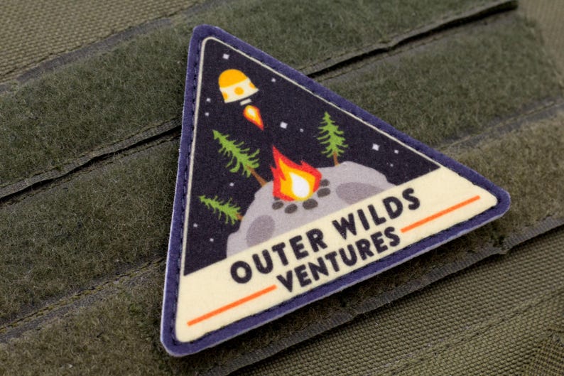 Może przedstawiać: Tr&oacute;jkątna naszywka z granatowym tłem przedstawiająca rakietę, ognisko i drzewa. Na naszywce widnieje napis "OUTER WILDS VENTURES" w białym kolorze z pomarańczowym podkreśleniem. Naszywka jest obramowana na niebiesko i leży na zielonej tkaninie.