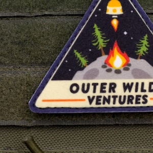 Może przedstawiać: Tr&oacute;jkątna naszywka z motywem kosmicznym i kempingowym. Naszywka przedstawia ognisko, drzewa, rakietę oraz tekst "OUTER WILDS VENTURES" pogrubioną czcionką. Naszywka jest obramowana na niebiesko i umieszczona na zielonym tle.