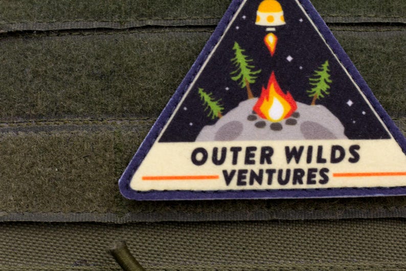 Może przedstawiać: Tr&oacute;jkątna naszywka z motywem kosmicznym i kempingowym. Naszywka przedstawia ognisko, drzewa, rakietę oraz tekst "OUTER WILDS VENTURES" pogrubioną czcionką. Naszywka jest obramowana na niebiesko i umieszczona na zielonym tle.