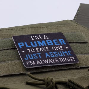 K&ouml;nnte beinhalten: Ein quadratischer Aufn&auml;her mit dem Text "I'M A PLUMBER TO SAVE TIME JUST ASSUME I'M ALWAYS RIGHT" in wei&szlig;er und blauer Schrift auf schwarzem Hintergrund. Der Aufn&auml;her ist an einer gr&uuml;nen taktischen Weste befestigt.