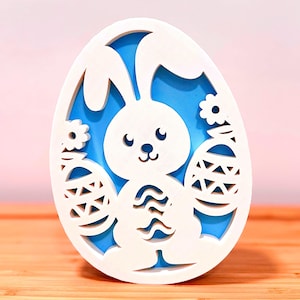 May include: White Easter egg decoration with a cutout design featuring a bunny and two decorated eggs, set against a blue backdrop. The decorative piece is on a wooden surface.