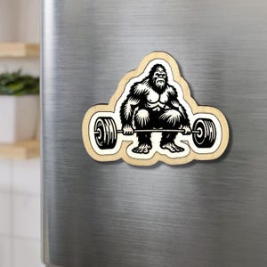 May include: A wooden magnet featuring a black and white illustration of a Bigfoot lifting a barbell. The Bigfoot is in a squat position, with the barbell in front of it. The magnet has a light brown border.