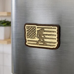 May include: A wooden magnet featuring a silhouette of Bigfoot walking across the American flag. The flag is made of wood and has a brown stain.