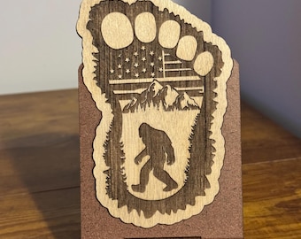 May include: A wooden phone stand shaped like a Bigfoot footprint with an American flag design and a silhouette of Bigfoot walking through a forest.
