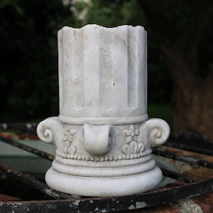 Könnte beinhalten: Ein weißer Beton-Pflanzkasten in Säulenform mit dekorativen Details. Der Pflanzkasten hat ein geriffeltes Design und eine Basis mit floralen Akzenten.