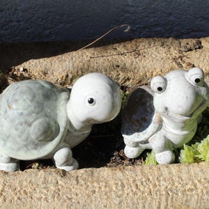 Könnte beinhalten: Zwei Keramik-Schildkrötenfiguren mit weißen Köpfen und graugrünen Panzern. Die Schildkröten sind auf einer Steinoberfläche mit kleinen grünen Pflanzen platziert. Eine Schildkröte blickt nach vorne, die andere ist zur Seite geneigt.
