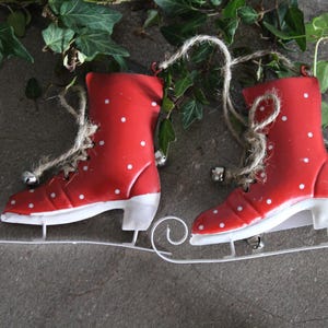 Puede incluir: Un par de adornos de patines de hielo rojos con lunares blancos y cuchillas blancas. Cada pat&iacute;n tiene una cuerda de yute para colgar y una peque&ntilde;a campana plateada. Los patines se colocan sobre un fondo gris con follaje verde.