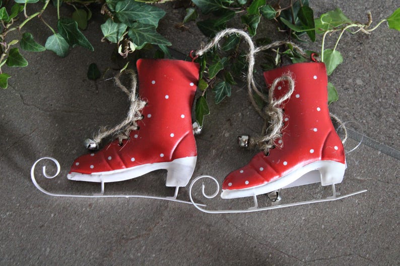 Puede incluir: Un par de adornos de patines de hielo rojos con lunares blancos y cuchillas blancas. Cada pat&iacute;n tiene una cuerda de yute para colgar y una peque&ntilde;a campana plateada. Los patines se colocan sobre un fondo gris con follaje verde.