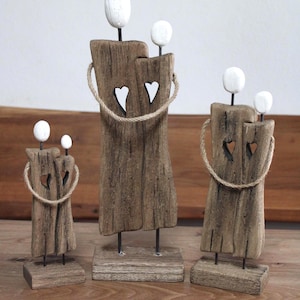Könnte beinhalten: Drei Holzskulpturen von Familien. Jede Skulptur hat einen Körper im Treibholz-Stil mit einem weißen Herz-Ausschnitt und einem Seildetail. Weiße, kugelförmige Köpfe sind oben an jeder Skulptur befestigt. Die Skulpturen sind auf Holzsockeln montiert.