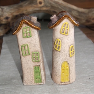 Könnte beinhalten: Zwei Keramikhausfiguren. Eine lehnt sich an, mit grünen Fenstern und Tür. Die andere ist aufrecht, mit gelben Fenstern und Tür. Beide haben braune Dächer und stehen auf neutralem Hintergrund.