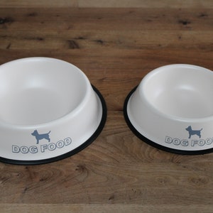 Könnte beinhalten: Zwei weiße Keramik-Hundenäpfe mit schwarzen Rändern. Die Näpfe haben eine graue Silhouette eines Hundes und den Text "DOG FOOD" auf dem Boden.