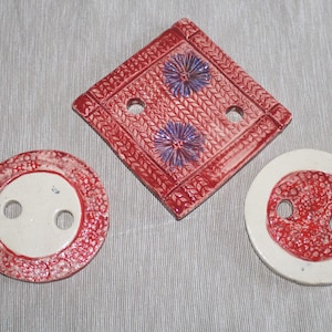 Könnte beinhalten: Drei handgefertigte Keramikteile: ein rotes und weißes rundes Stück, ein rotes rautenförmiges Stück und ein zweites rundes Stück. Das rautenförmige Stück hat zwei lila Blumendesigns und zwei Löcher. Die runden Stücke haben zwei Löcher und rot-weiße Muster.