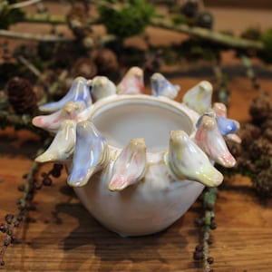 Könnte beinhalten: Ein weißer Keramik-Pflanztopf in Form eines Vogelrings. Die Vögel sind in Blautönen, Rosa und Gelb bemalt. Der Topf ist perfekt für kleine Pflanzen oder Sukkulenten.