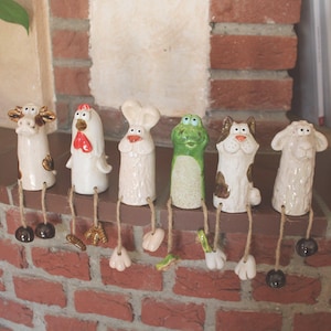 Könnte beinhalten: Eine Sammlung von sechs Keramik-Tierfiguren, darunter eine Kuh, ein Huhn, ein Koch, ein Frosch, ein Hund und ein Schaf. Jede Figur hat hängende Beine und ist in verschiedenen Farben bemalt. Sie sind auf einem Ziegelsims angeordnet.