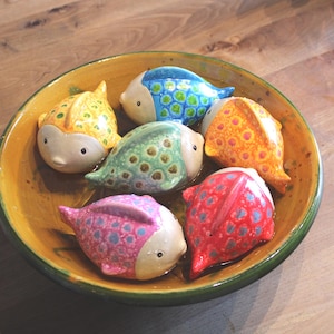Könnte beinhalten: Eine Keramikschale mit gelbem Innenraum und grünem Rand enthält sechs bunte Fischfiguren. Die Fische sind in verschiedenen Farben bemalt, darunter Gelb, Blau, Grün, Orange, Rosa und Rot, mit gemusterten Details. Die Schale steht auf einer Holzoberfläche.