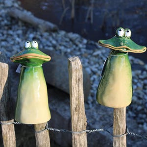 Könnte beinhalten: Zwei Keramik-Froschfiguren in Grün und Weiß bemalt, die auf Holzpfosten eines Zauns sitzen. Die Frösche haben große Augen und schauen nach vorne.