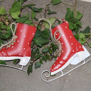 Puede incluir: Dos patines de hielo decorativos rojos con lunares blancos y cuchillas blancas. Cada pat&iacute;n tiene una peque&ntilde;a campana plateada y est&aacute; atado con hilo. La hiedra verde rodea los patines.