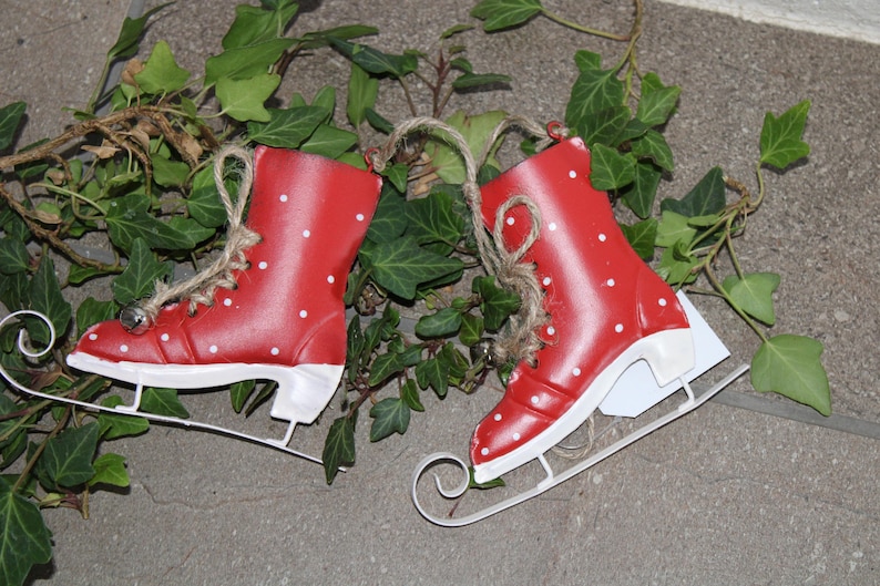 Puede incluir: Dos patines de hielo decorativos rojos con lunares blancos y cuchillas blancas. Cada pat&iacute;n tiene una peque&ntilde;a campana plateada y est&aacute; atado con hilo. La hiedra verde rodea los patines.