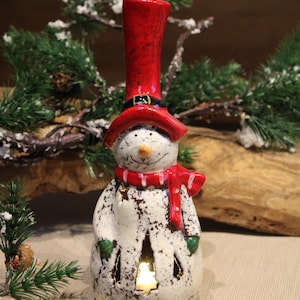 Könnte beinhalten: Keramik-Schneemannfigur mit rotem Zylinder, Schal und grünen Fäustlingen. Der Schneemann hat ein lächelndes Gesicht und ausgeschnittene Formen, durch die Licht scheint. Eine festliche Weihnachtsdekoration.