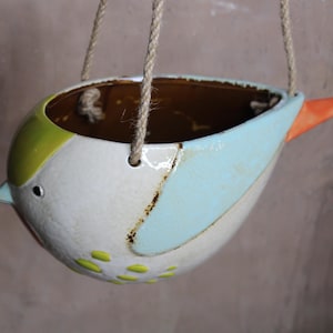 Könnte beinhalten: Ein weißer Keramik-Hängepflanztopf in Form eines Vogels mit einem hellblauen Flügel, grünen Akzenten und einem orangefarbenen Schnabel.