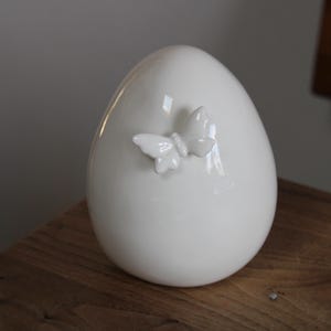 Könnte beinhalten: Weiße Keramikvase in Eiform mit einem zarten Schmetterlingsdesign.