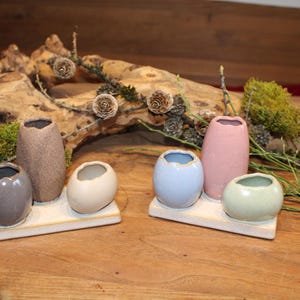 Könnte beinhalten: Ein Set aus sechs kleinen Keramikvasen in verschiedenen Farben, darunter Grau, Beige, Blau, Rosa und Grün, angeordnet auf zwei rechteckigen weißen Tabletts. Die Vasen haben einzigartige Formen und Texturen, ideal für kleine Blumen.