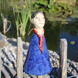 Könnte beinhalten: Keramik-Gartendekoration, die eine Figur mit blauem Gewand, rotem Schal und neutralem Hautton darstellt. Die Figur ist auf einem Holzzaun montiert, mit einem Teich und Grün im Hintergrund.