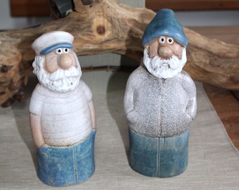 Puede incluir: Dos figuras de gnomos de cerámica. Uno lleva un gorro de marinero blanco y una camisa a rayas, el otro un gorro azul y una chaqueta gris. Ambos tienen barbas blancas y pantalones azules. Se exhiben sobre una superficie beige.