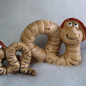 Könnte beinhalten: Zwei Keramik-Wurmfiguren, eine größer als die andere. Der größere Wurm ist braun mit einem roten Hut und einem lächelnden Gesicht. Der kleinere Wurm ist braun mit einem roten Hut und einem lächelnden Gesicht.