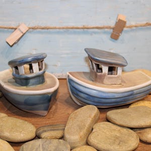 Könnte beinhalten: Zwei Keramik-Bootsfiguren mit blauen und weißen Details, jeweils mit Kabinen. Sie sind von glatten Kieselsteinen umgeben, mit einem Seil und Wäscheklammern im Hintergrund. Das nautische Thema erinnert an eine Küsten- oder Meereslandschaft.