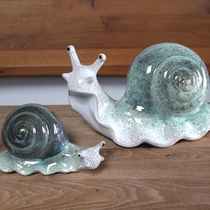 Könnte beinhalten: Zwei Schneckenfiguren aus Keramik mit glänzender Oberfläche. Die größere Schnecke hat ein hellgrünes Gehäuse und einen weißen Körper mit strukturierten Punkten. Die kleinere Schnecke hat ein dunkelblau-graues Gehäuse und einen hellgrünen Körper.