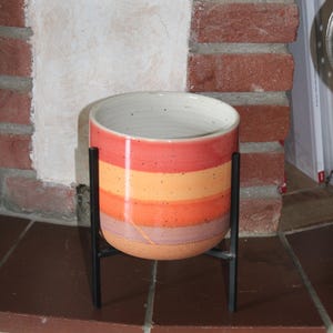 Könnte beinhalten: Ein Keramik-Pflanztopf mit einem gestreiften Design in Rot-, Orange- und Pfirsichtönen, auf einem schwarzen Metallständer. Der Pflanztopf hat eine zylindrische Form und einen weißen Innenraum. Der Ständer hat ein minimalistisches Design.