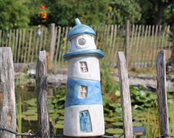 Puede incluir: Una figura de faro de cerámica con un patrón de rayas blancas y azules, un techo cónico y pequeñas ventanas y una puerta empotradas. La estructura es cilíndrica con un acabado liso y se asienta sobre un poste de madera al aire libre.